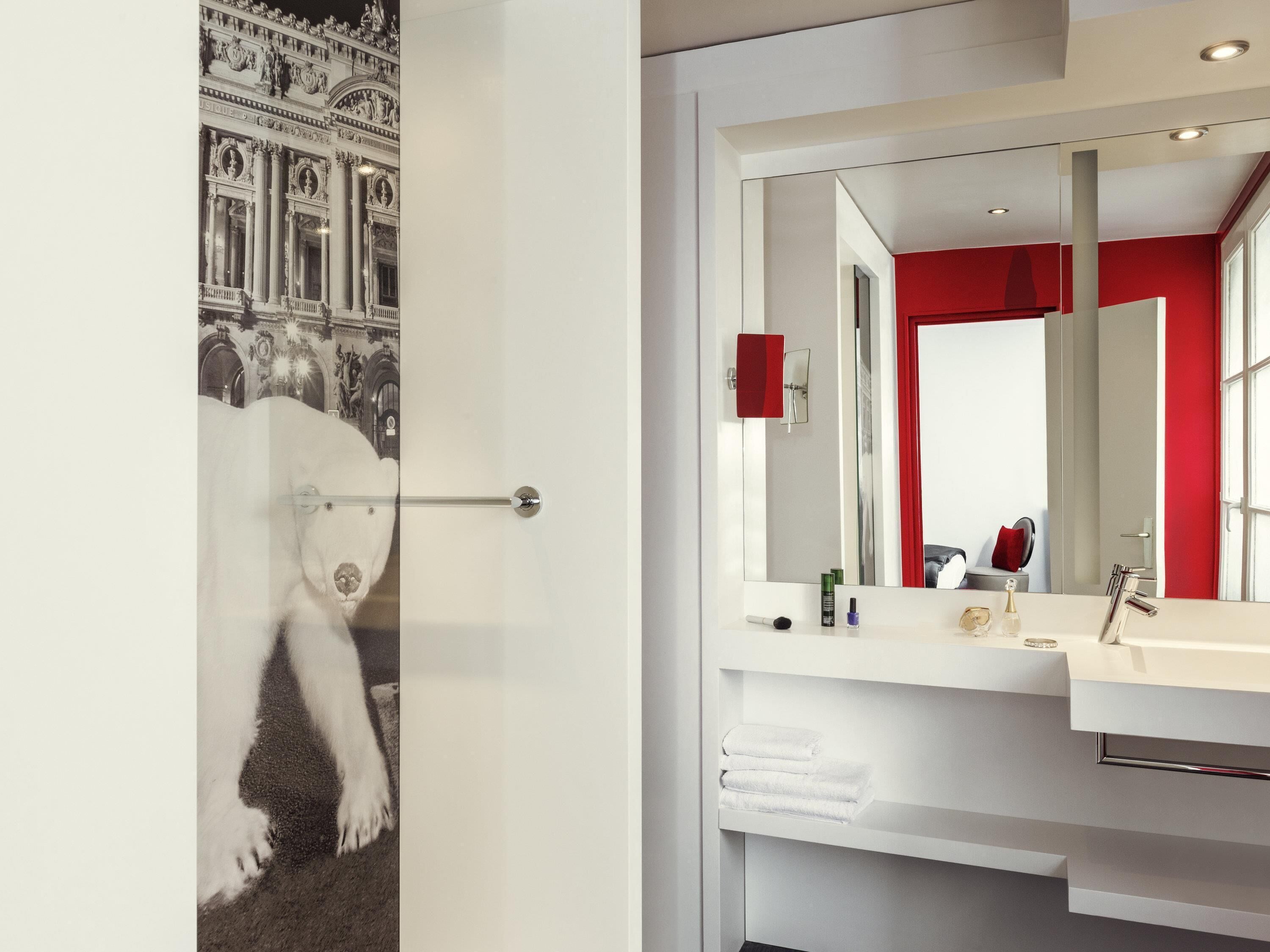 Um banheiro moderno com um espelho grande, uma parede de destaque vermelha e uma porta com fotografia de arquitetura clássica.