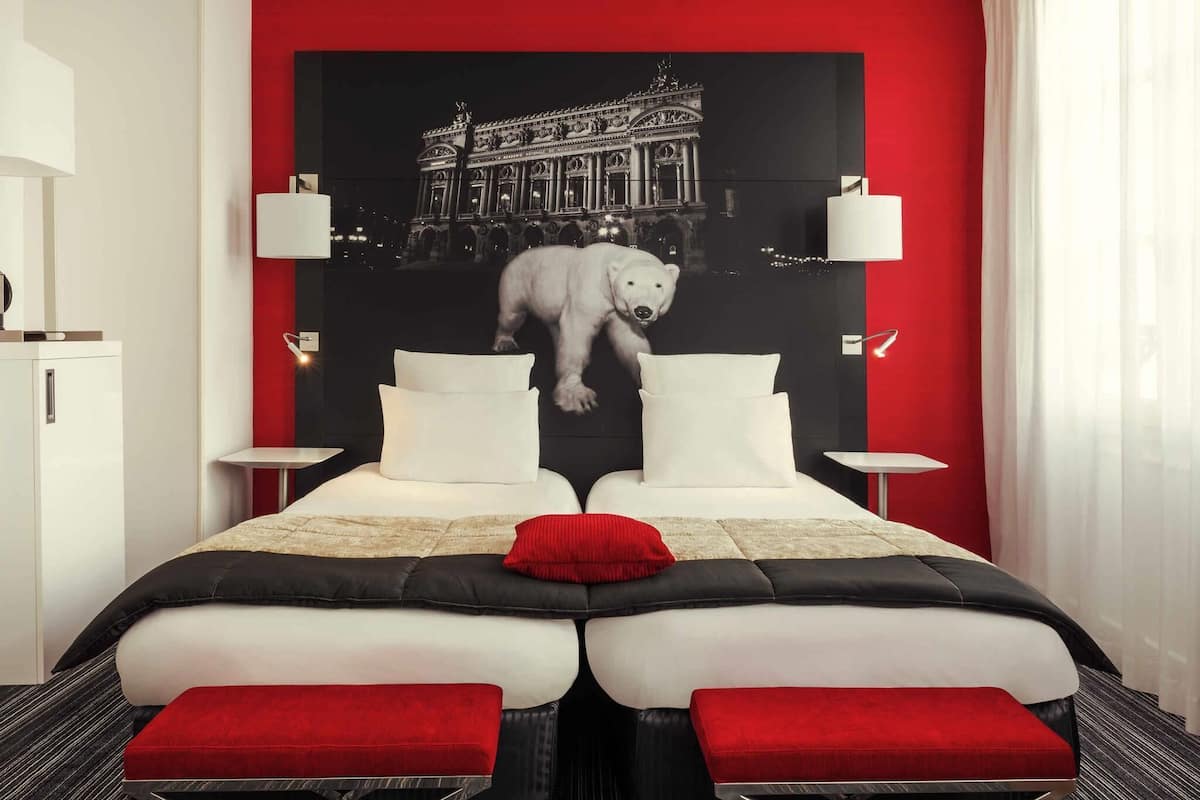 A hotel room with a bed, two bedside tables, a red accent wall, and a large poster of a polar bear and a building.