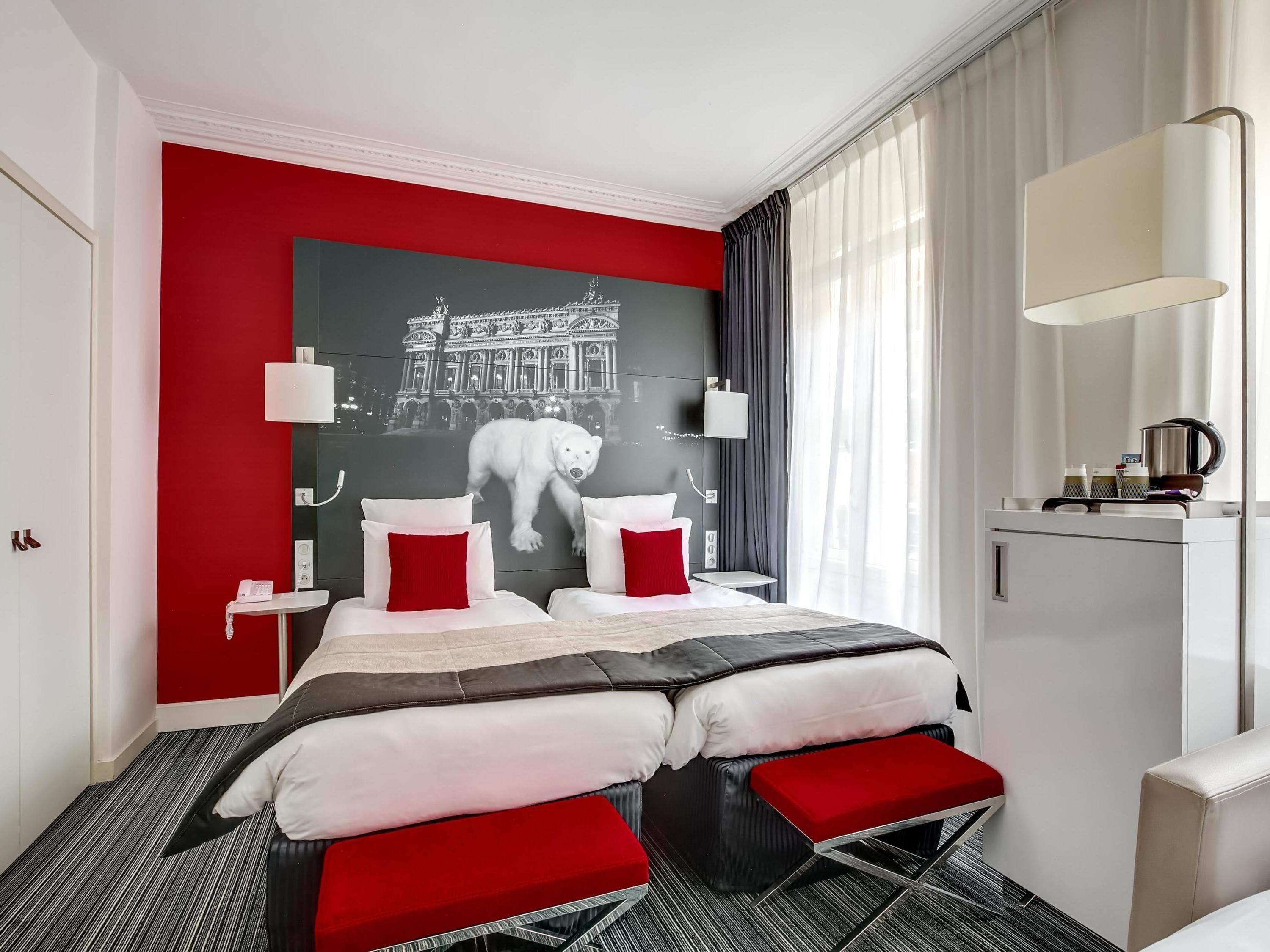 Une chambre d’hôtel avec un mur d’accent rouge, un lit avec une literie blanche et rouge, une petite table de chevet avec une lampe, et une vue sur un immeuble par la fenêtre.