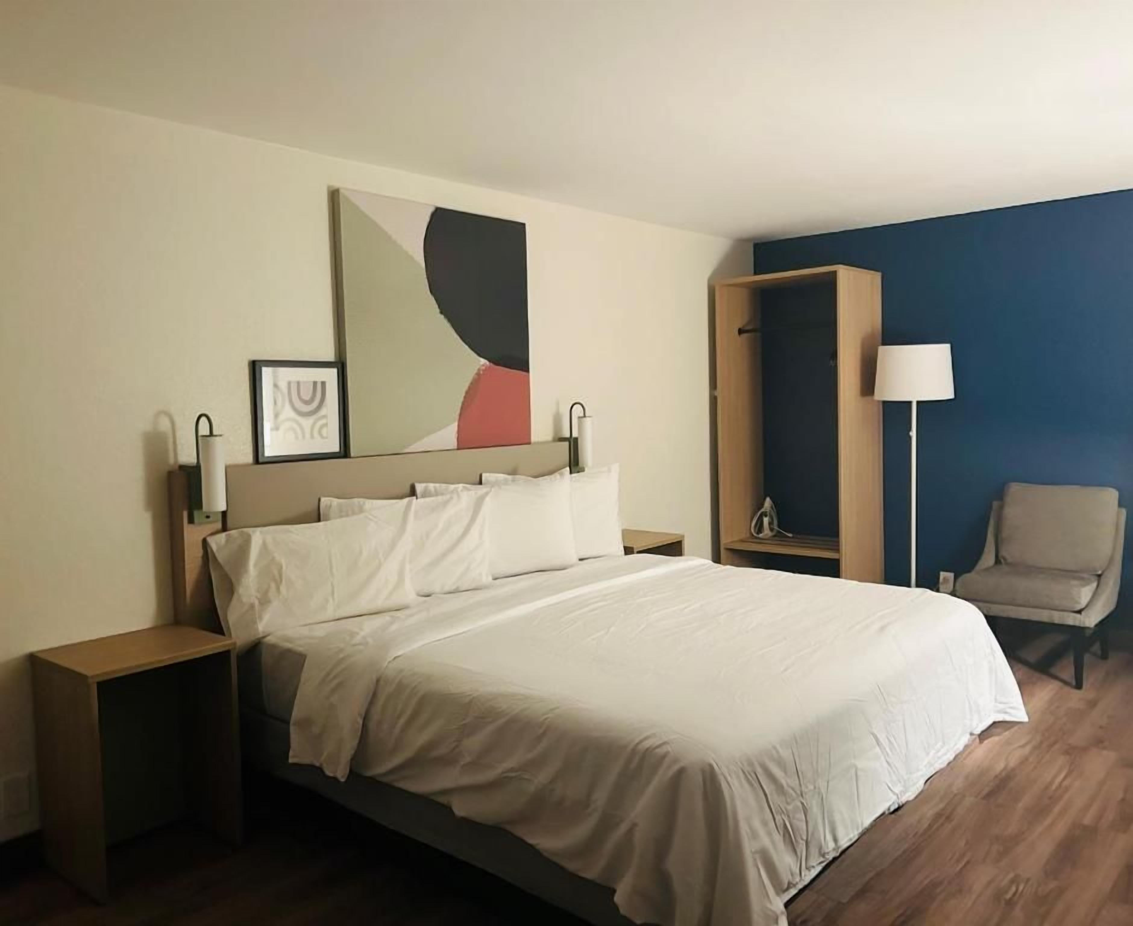 Quarto de hotel com cama, mesas de cabeceira, uma cadeira, abajur e um quadro na parede.