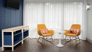 Una habitación moderna con una consola de madera, dos mecedoras con cojines coloridos, una mesita redonda y un ventanal con cortinas de gasa.