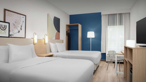 Habitación de hotel con dos camas, un escritorio, una silla, un televisor y obras de arte en la pared.
