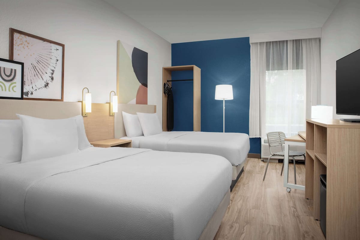 Habitación de hotel con dos camas, un escritorio, una silla, un televisor y obras de arte en la pared.