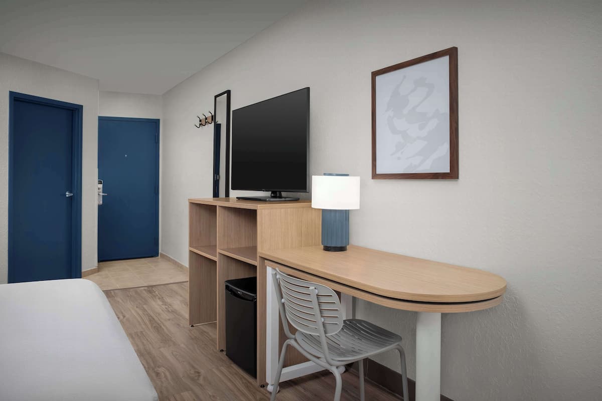 Habitación de hotel con cama, escritorio con silla, televisor y un cuadro en la pared.