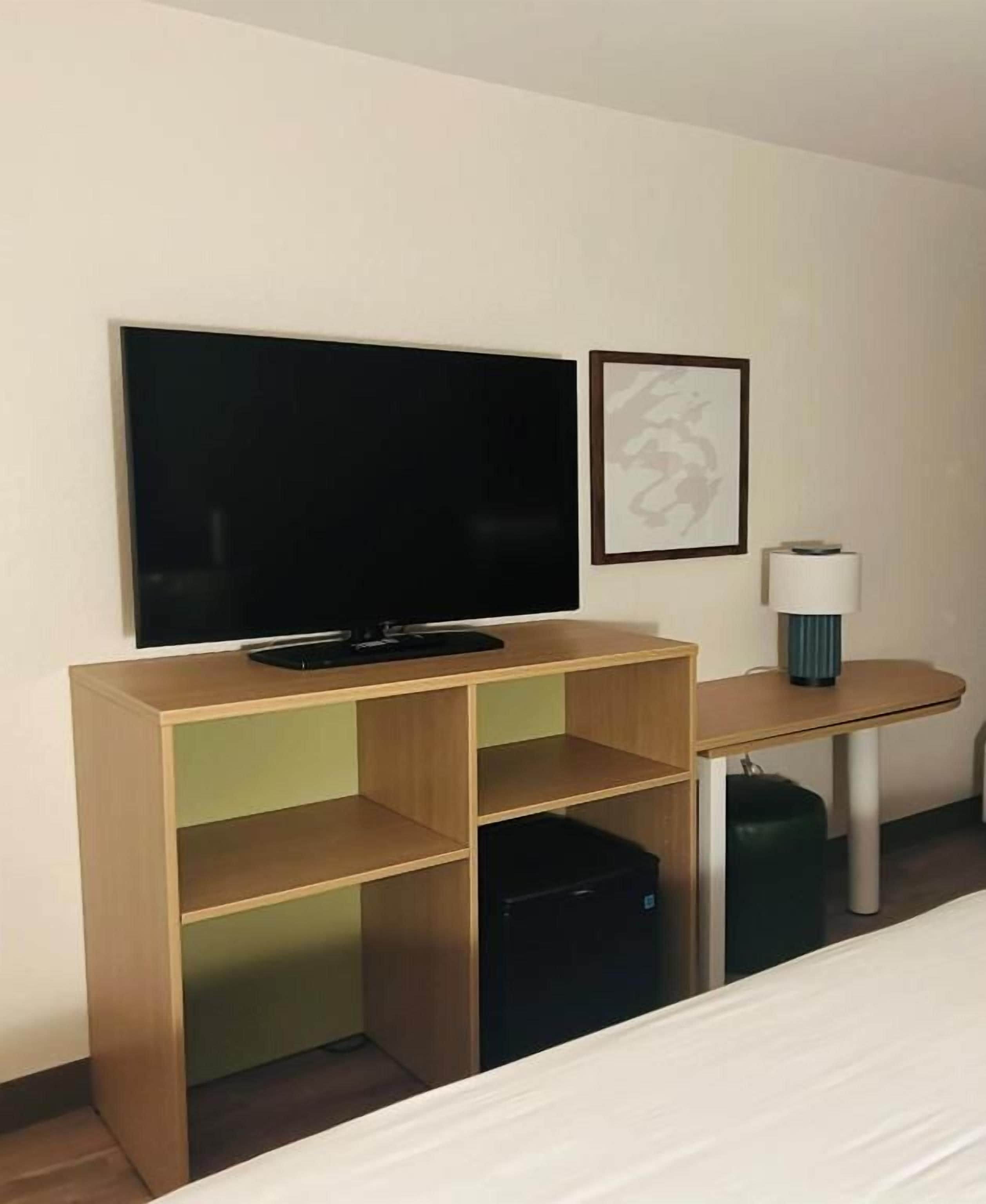 A hotel room with a bed, a TV mounted on a wooden shelf, a small table with a lamp, and a trash bin.