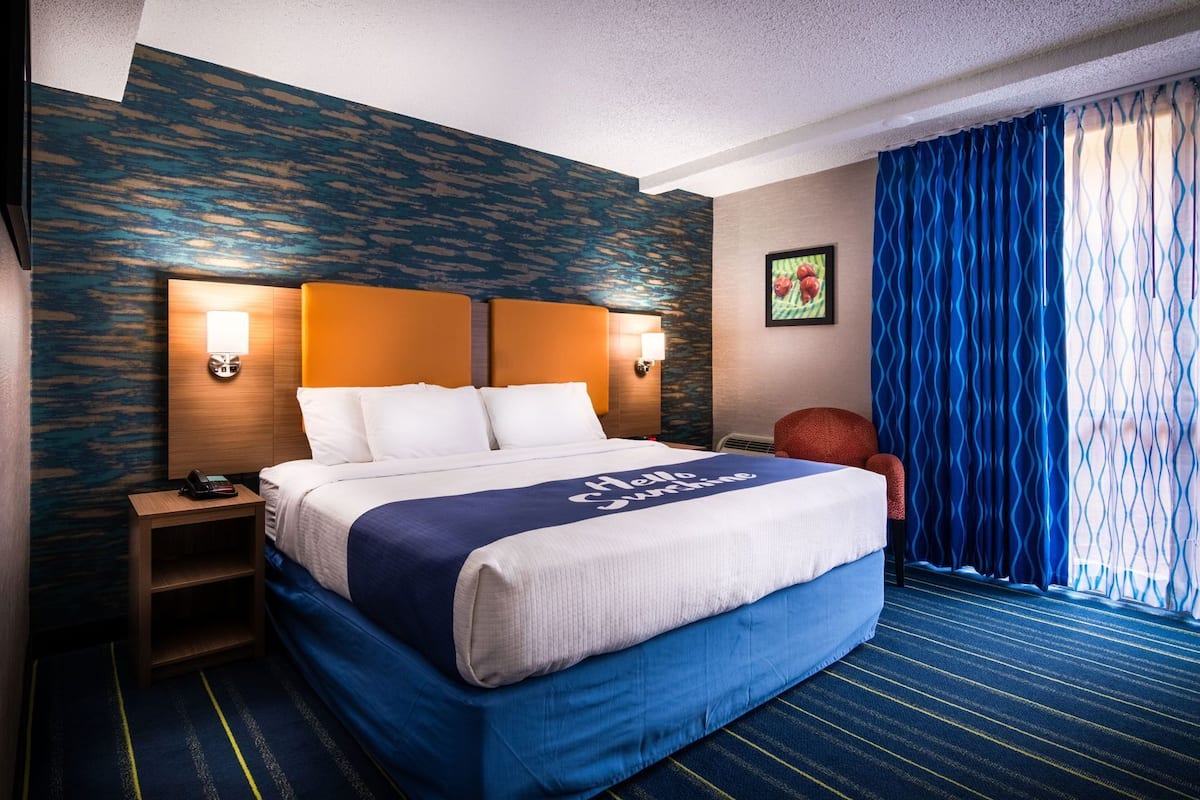 A hotel room with a large bed, two bedside lamps, a chair, a small table, and a window with blue curtains.