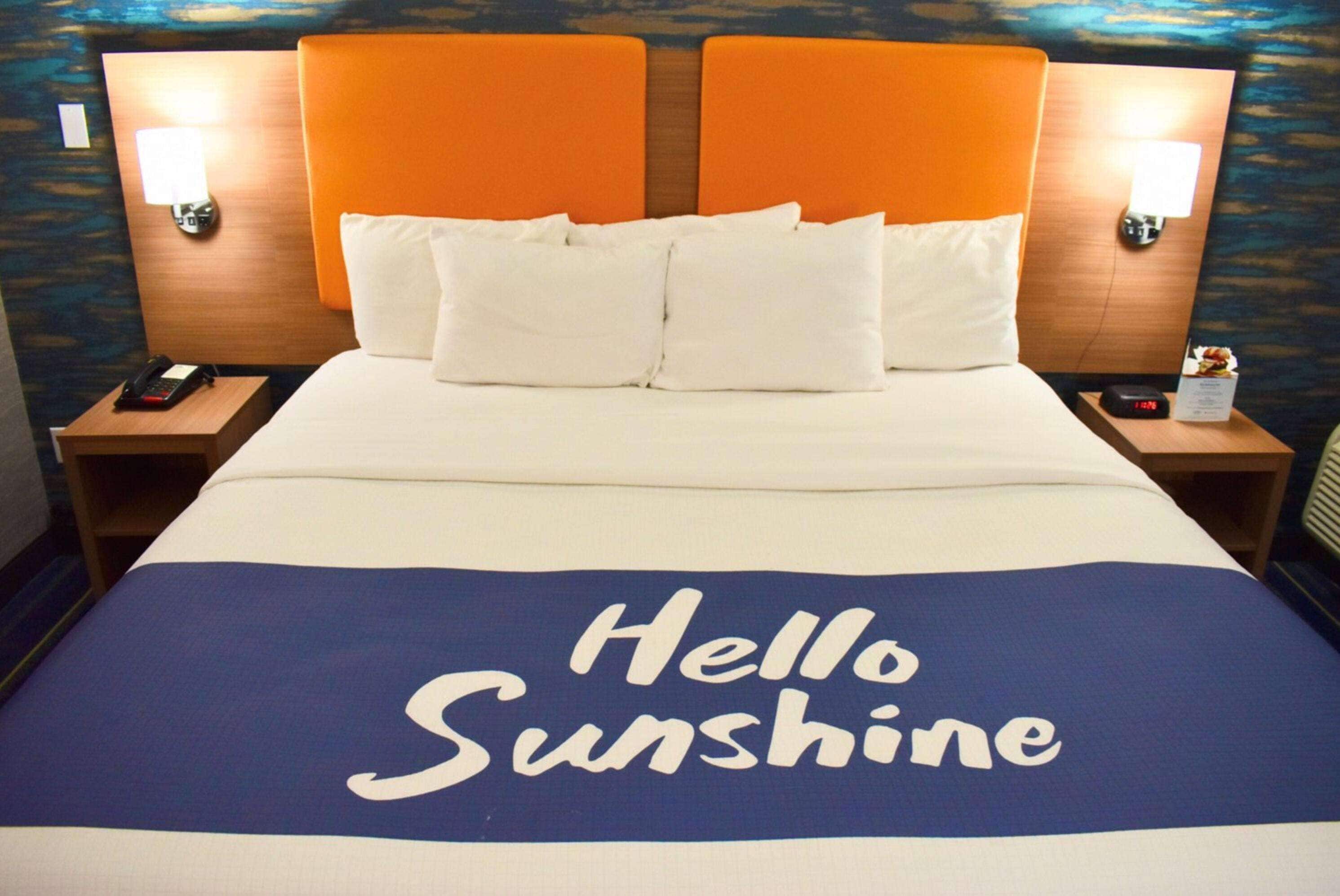A hotel room with a bed featuring a 'Hello Sunshine' bedspread, two nightstands, a phone, and a clock.