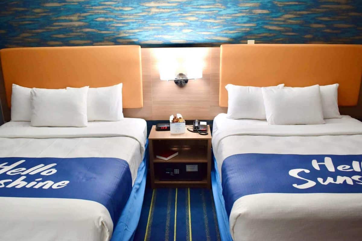 A hotel room with two beds, each with a blue bedspread and white pillows, a nightstand with a lamp, a clock, and a phone.