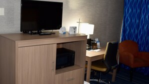 Una habitación de hotel con escritorio de madera, televisión, microondas, lámpara, silla y un cesto de basura.