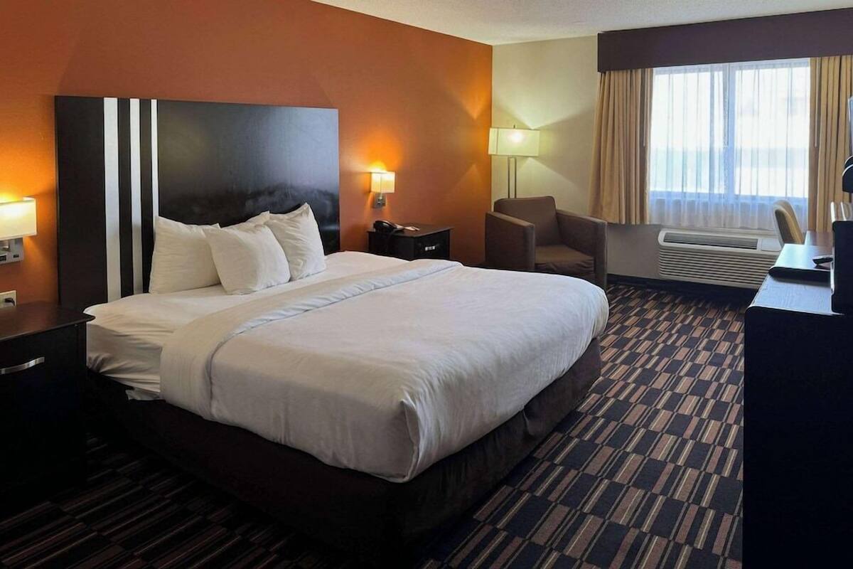 A hotel room with a large bed, a desk, a chair, a lamp, and an air conditioning unit.