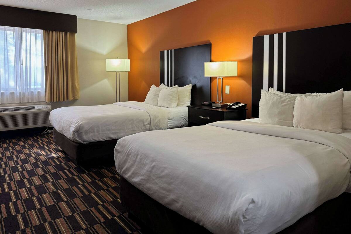 A hotel room with two beds, a large window with curtains, a wall-mounted air conditioner, and two bedside lamps.