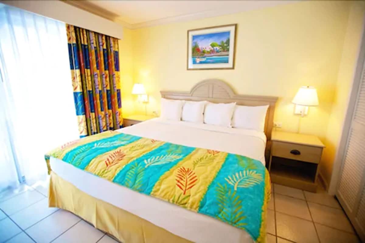 A hotel room with a large bed, two bedside tables with lamps, a window with curtains, and a painting on the wall.