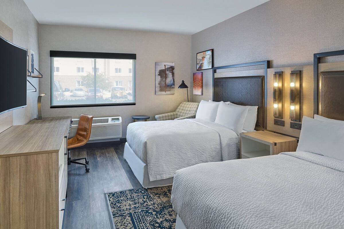Ein Hotelzimmer mit zwei Betten, einem Schreibtisch, einem Stuhl, einem Fernseher und einem großen Fenster mit Blick auf die Stadt.