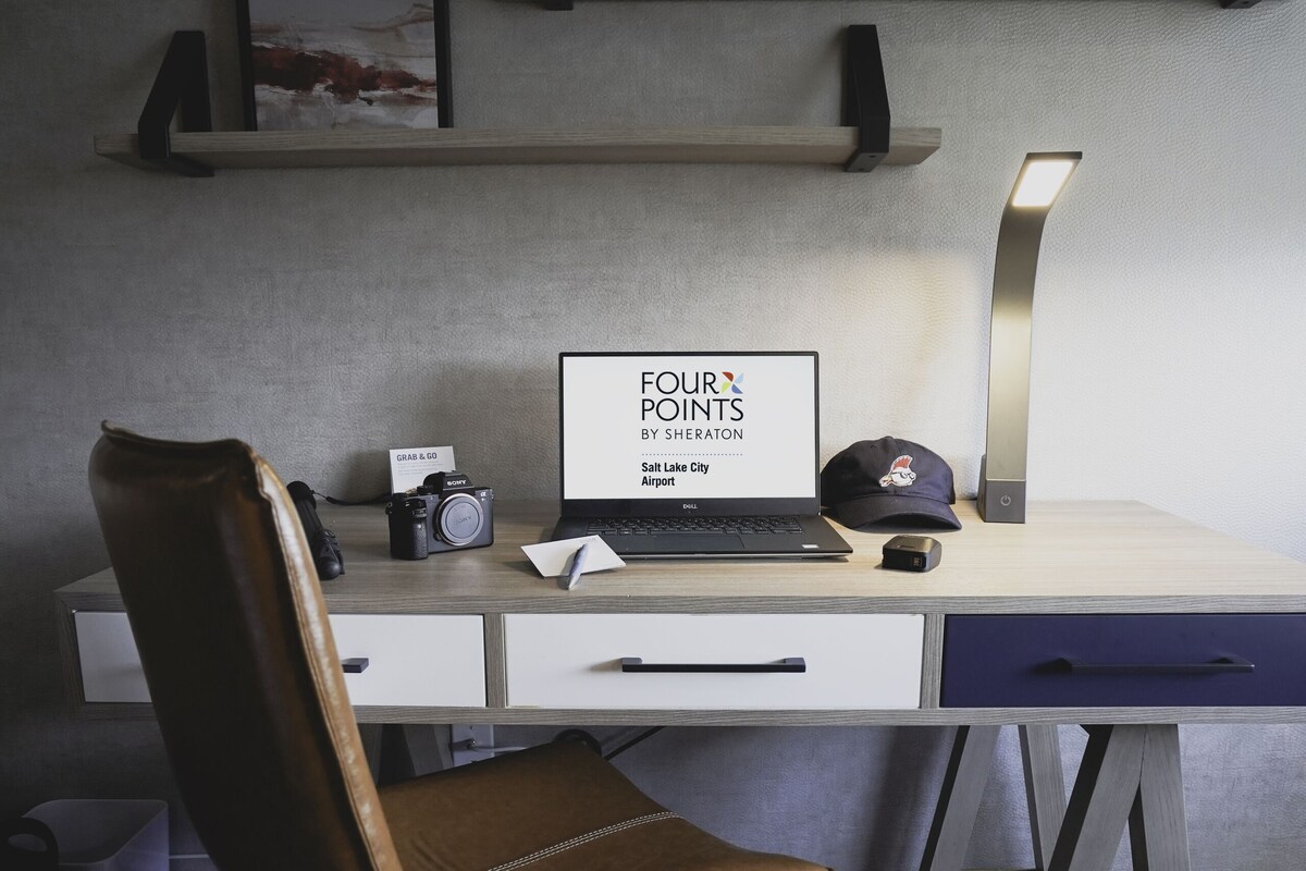 A modern office desk with a laptop, camera, and cap, a leather chair, and artwork on shelves.