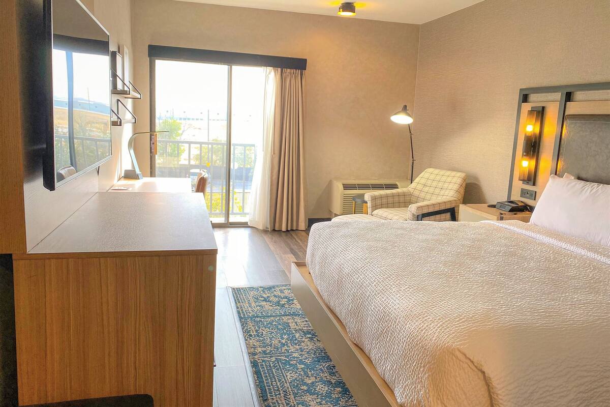 Ein Hotelzimmer mit einem großen Bett, einem Schreibtisch, einem Sessel, einem Flachbildfernseher und einem Balkon mit Vorhängen.