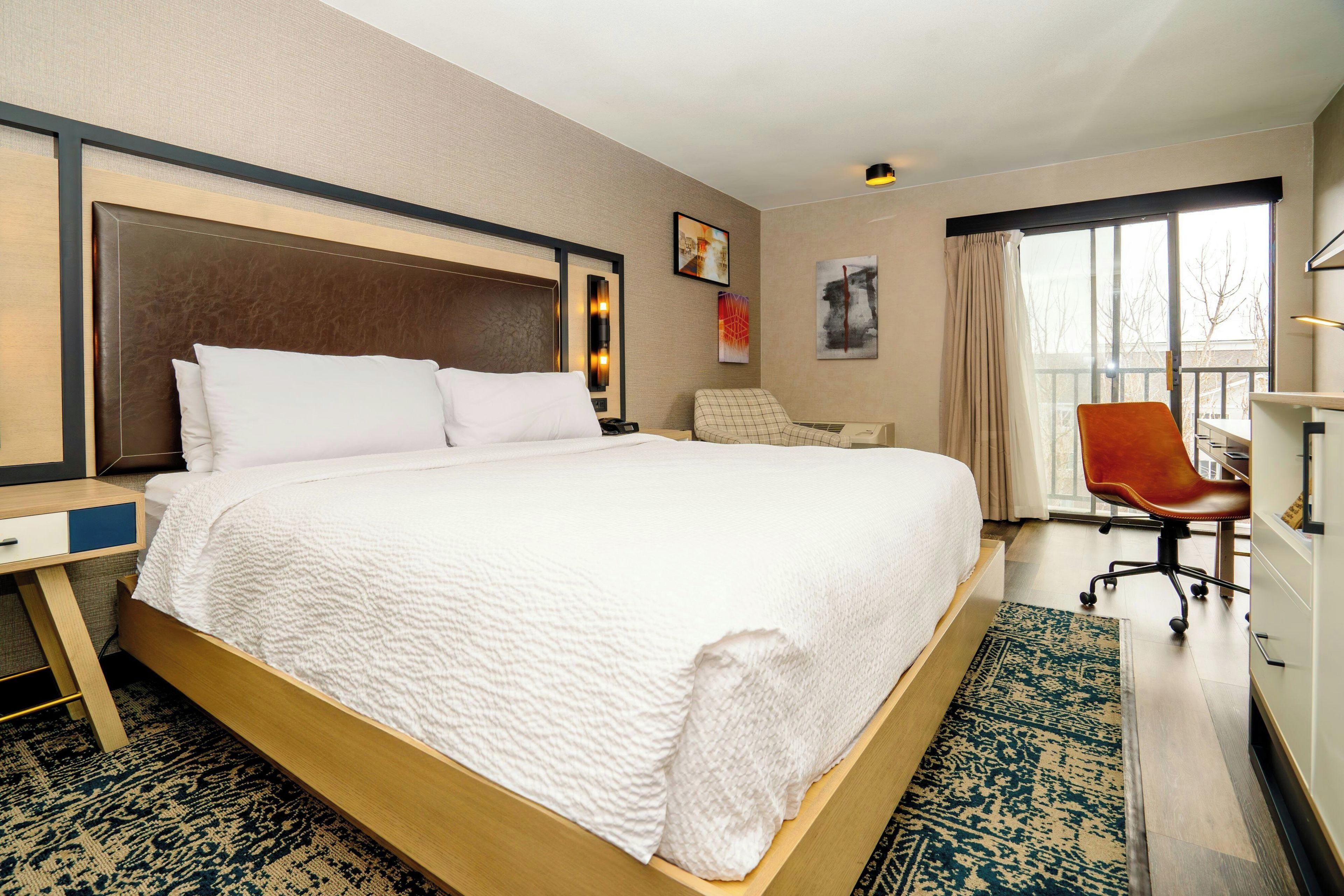 Une chambre d’hôtel avec un grand lit, un bureau, une chaise, une fenêtre avec des rideaux et des œuvres d’art accrochées aux murs.
