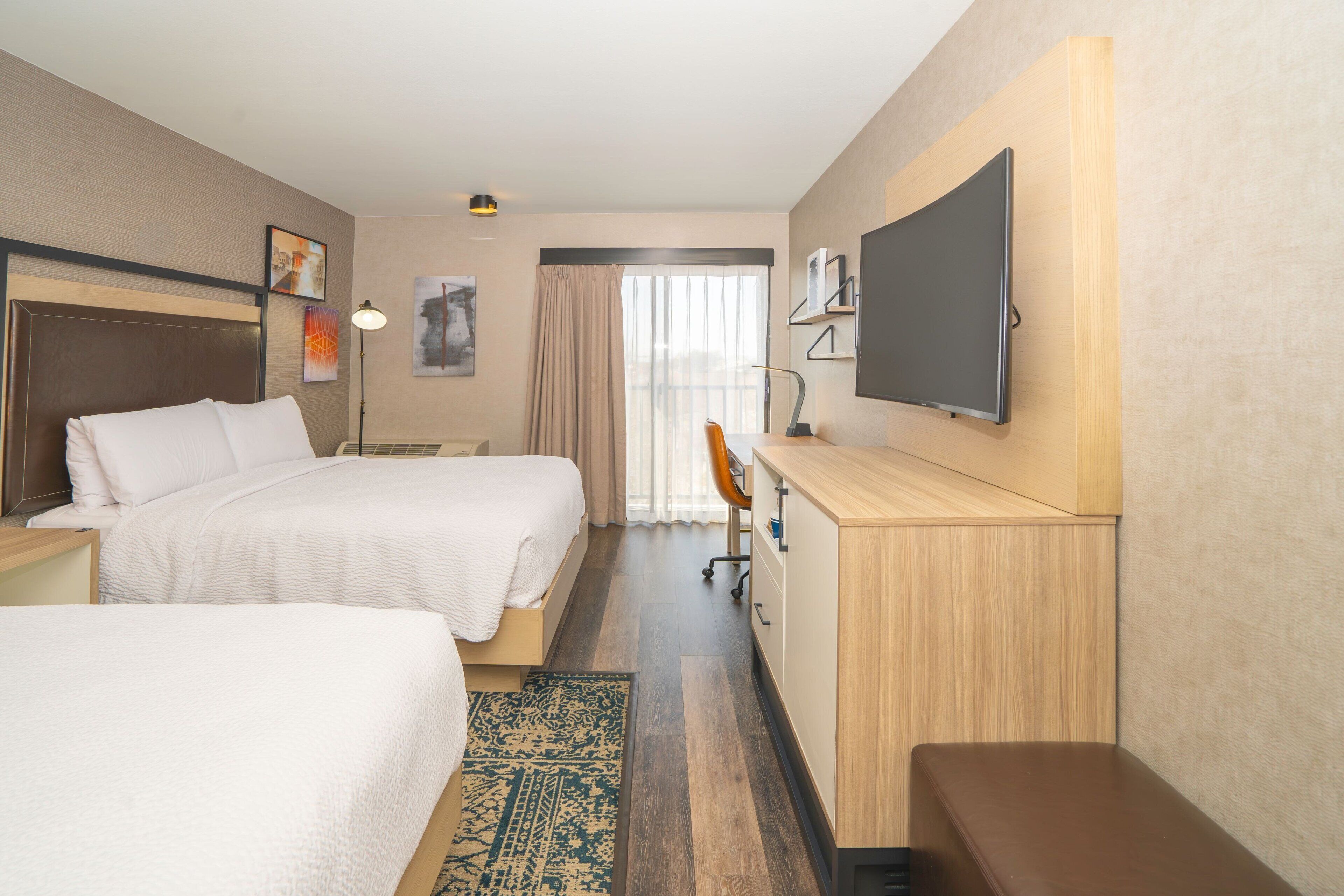 A hotel room with two beds, a desk, a TV, and a window with curtains.