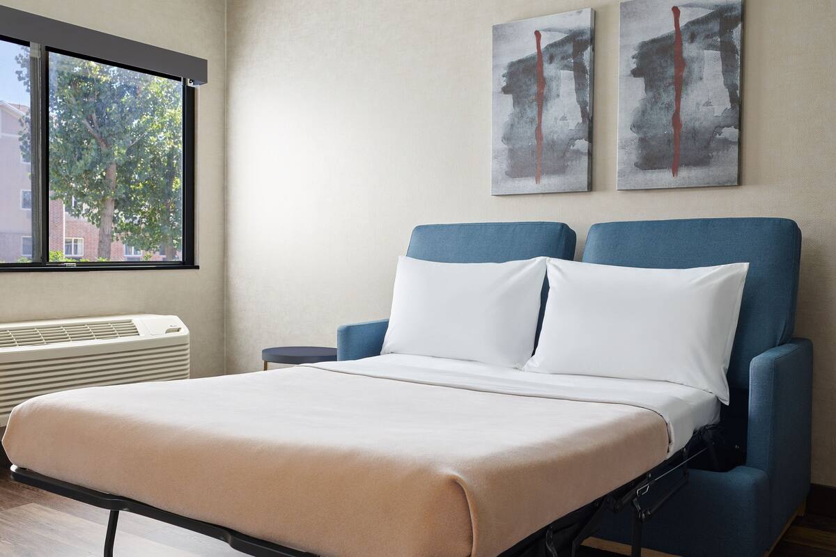 Ein ordentlich bezogenes Bett mit weißer Matratze und blauem Kopfteil, ein Fenster mit Blick auf Bäume und Gebäude sowie zwei abstrakte Gemälde an der Wand.
