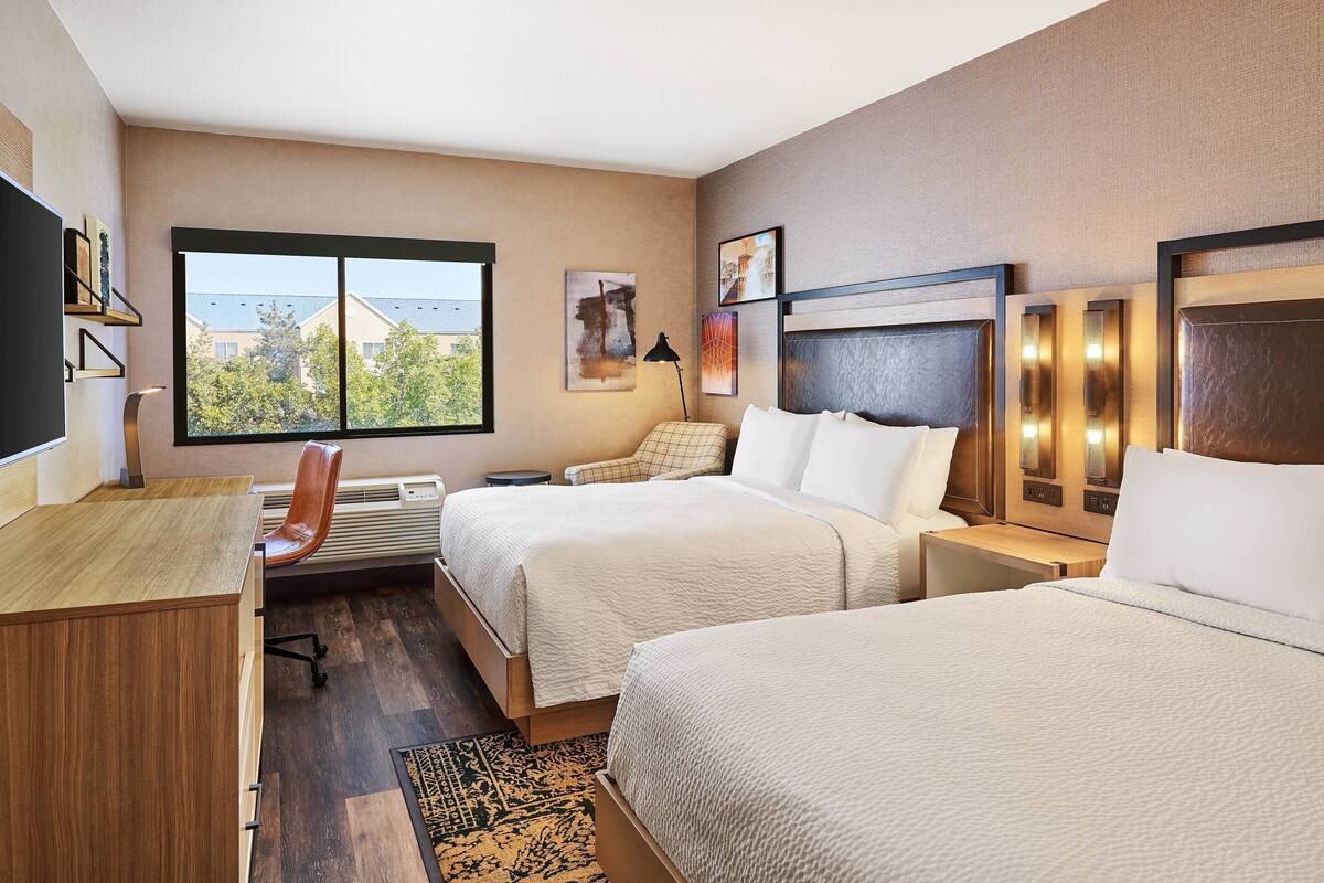 Ein Hotelzimmer mit zwei Betten, einem Schreibtisch und einem Sessel. Ein großes Fenster bietet Blick auf Gebäude und Bäume.