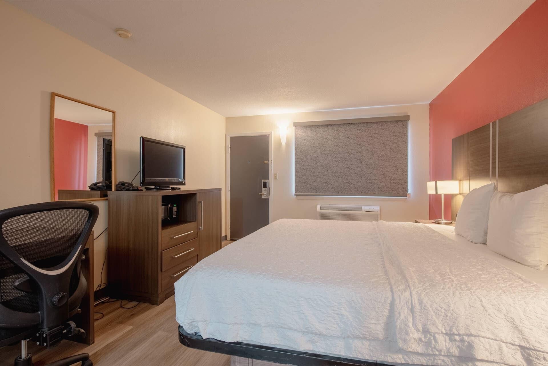 Habitación de hotel con cama, escritorio y silla, televisor, espejo y ventana con persiana.