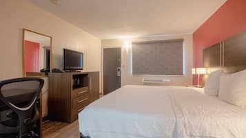 Habitación de hotel con cama, escritorio y silla, televisor, espejo y ventana con persiana.