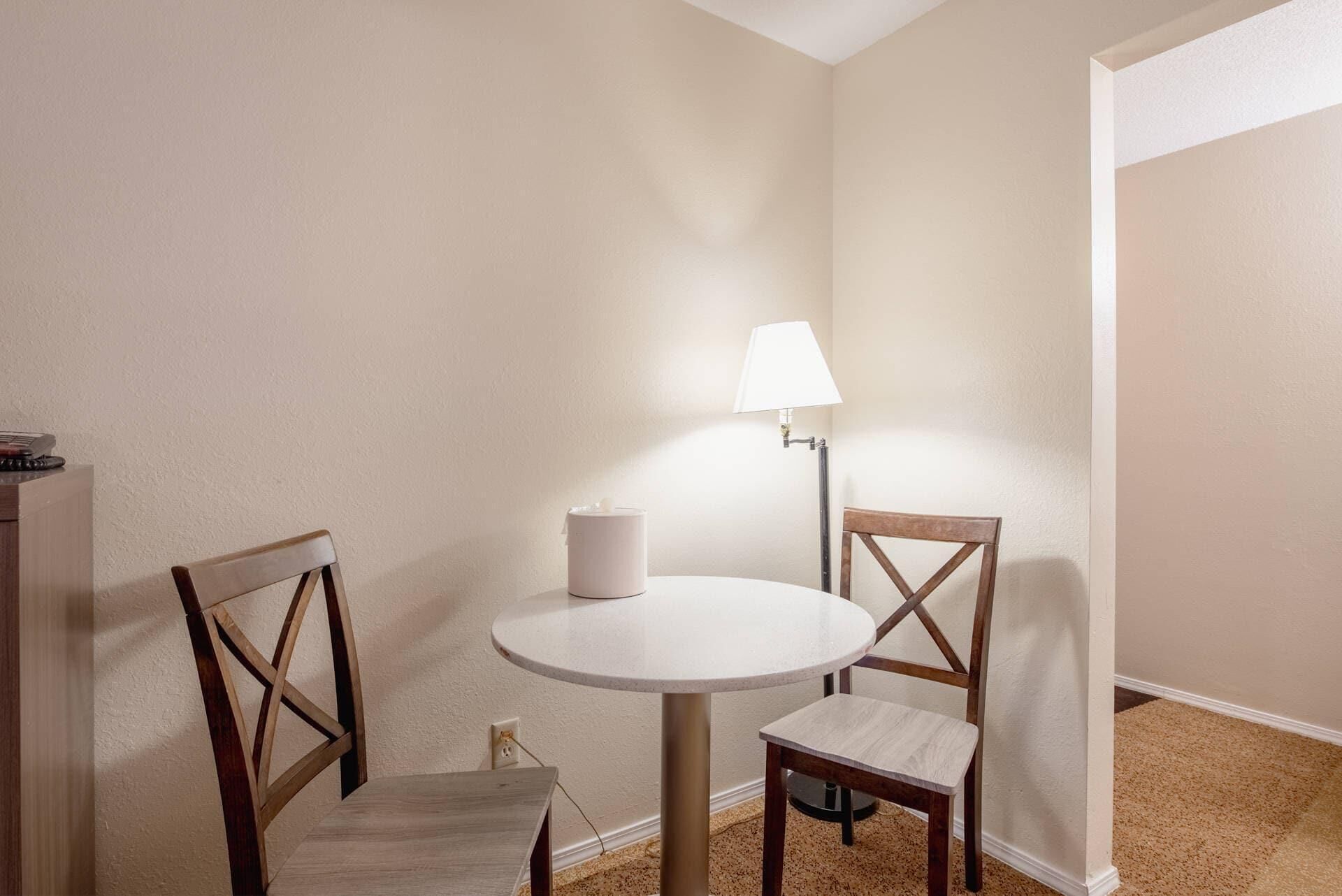 Una pequeña mesa redonda de comedor con dos sillas de madera, una lámpara blanca y un recipiente blanco sobre la mesa.