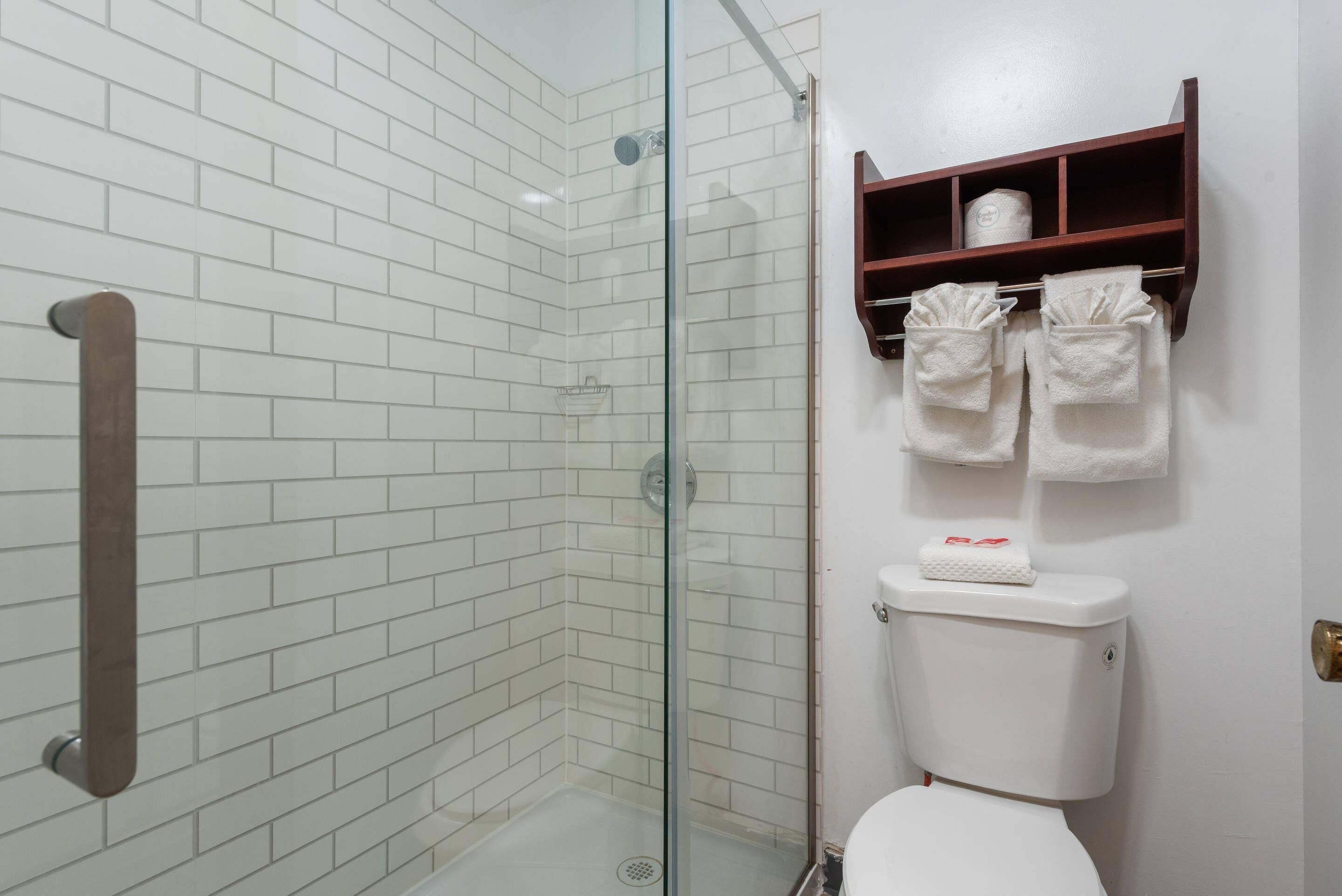 Un baño con paredes de azulejos blancos, cabina de ducha con mampara de vidrio, toallero de madera y un inodoro con estante superior.