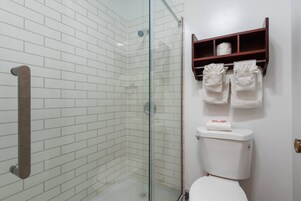 Un baño con paredes de azulejos blancos, cabina de ducha con mampara de vidrio, toallero de madera y un inodoro con estante superior.
