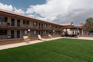 Un motel de varios pisos con techo plano, patio cubierto de césped y mesas al aire libre.