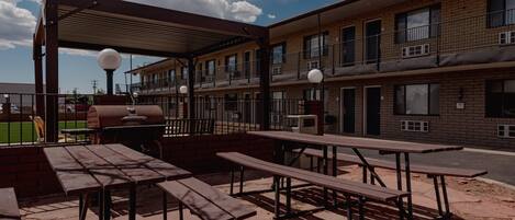 Un patio al aire libre con mesas de picnic y una parrilla, junto a un edificio de varios pisos con balcones.