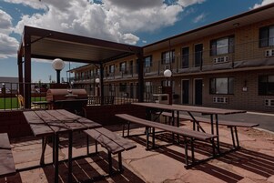 Un patio al aire libre con mesas de picnic y una parrilla, junto a un edificio de varios pisos con balcones.
