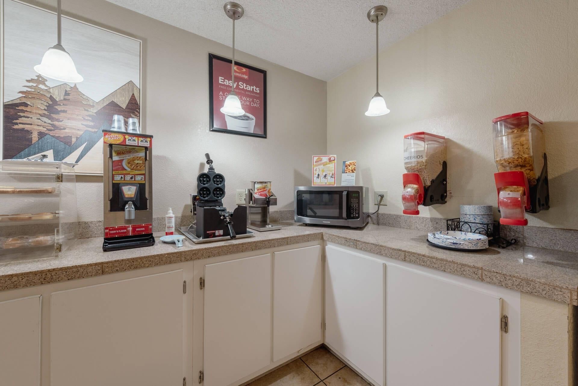 A kitchenette with a microwave, toaster, and coffee machine. There are cereal boxes and a poster on the wall.