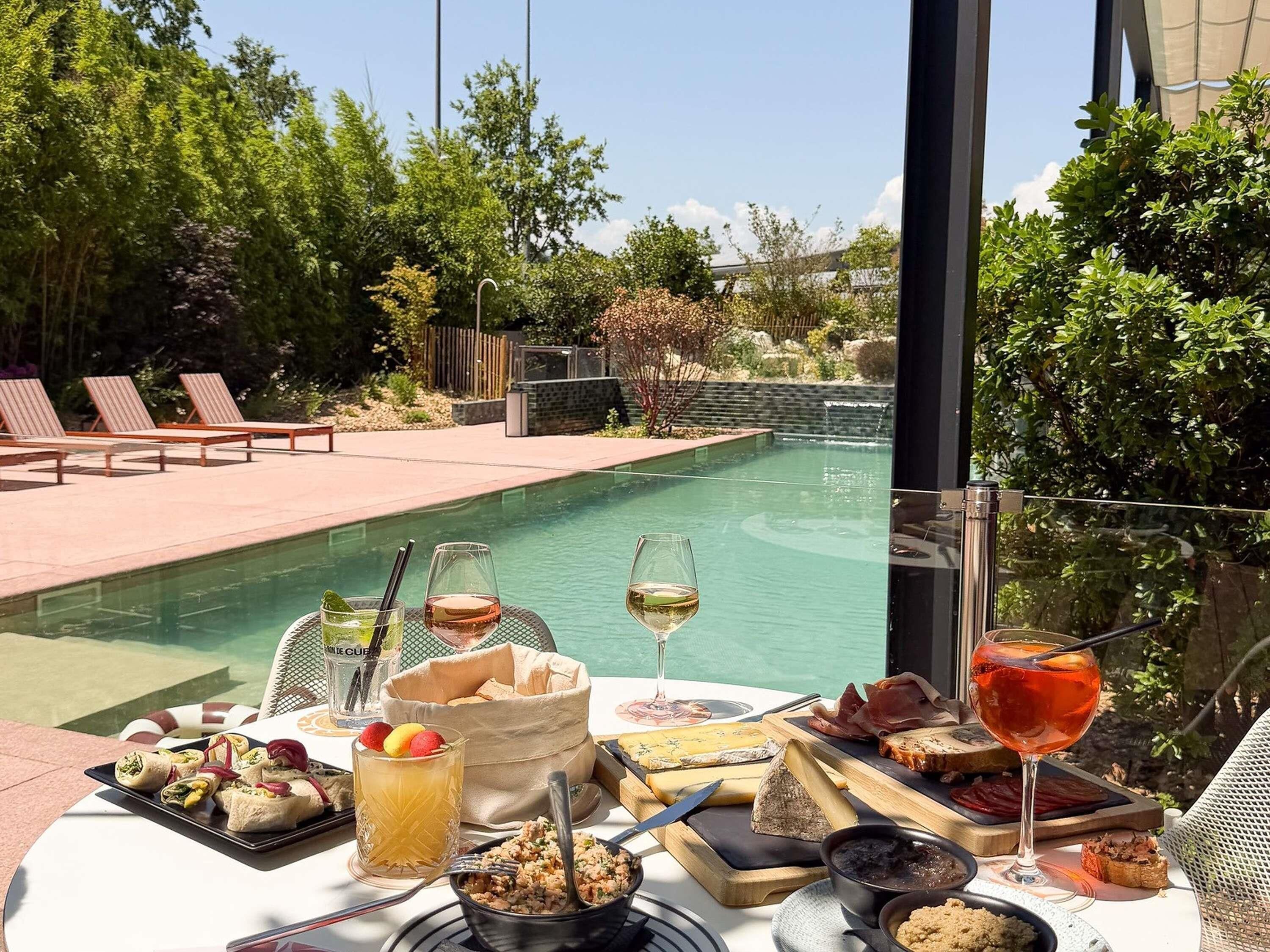 Un espace repas aménagé au bord de la piscine, avec une table dressée pour un repas, comprenant divers fromages, de la charcuterie et une bouteille de vin.
