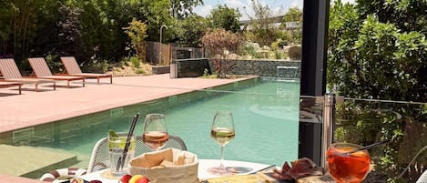 A poolside dining area with a table set for a meal, including various cheeses, meats, and a glass of wine.