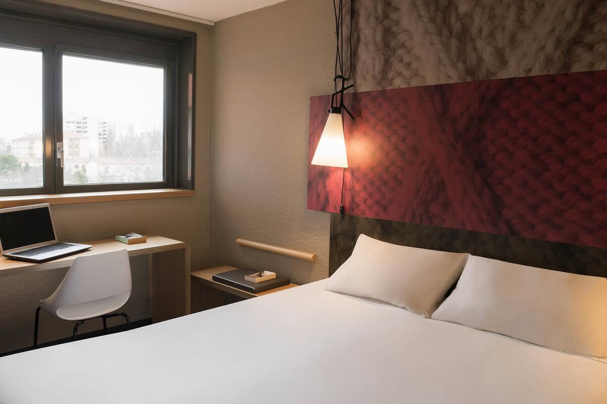 Une chambre d’hôtel avec un lit, un bureau avec un ordinateur portable, une chaise et une fenêtre donnant sur des immeubles.