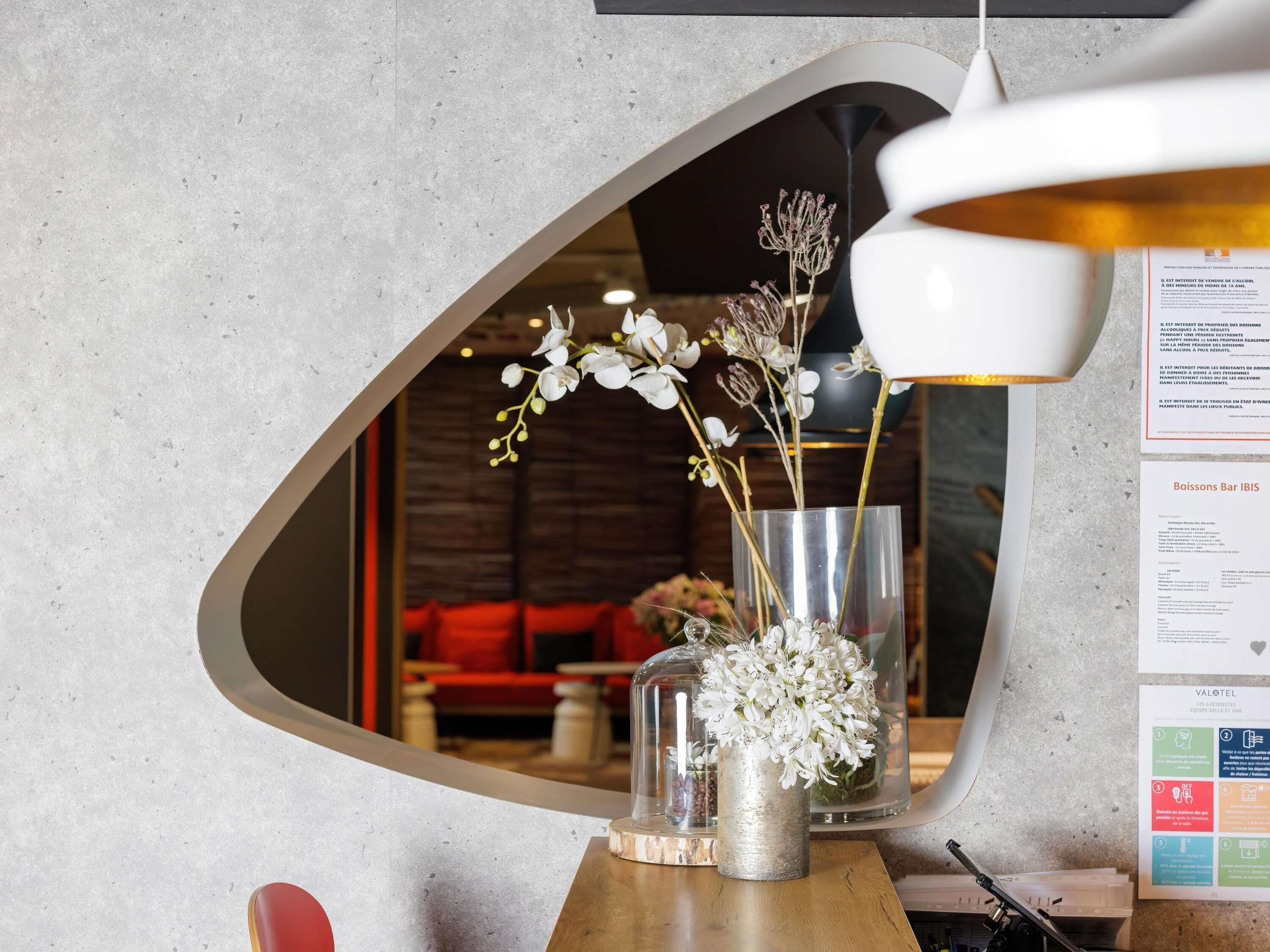 A mirror reflecting a room with a red couch, a glass vase with flowers, and a menu board.