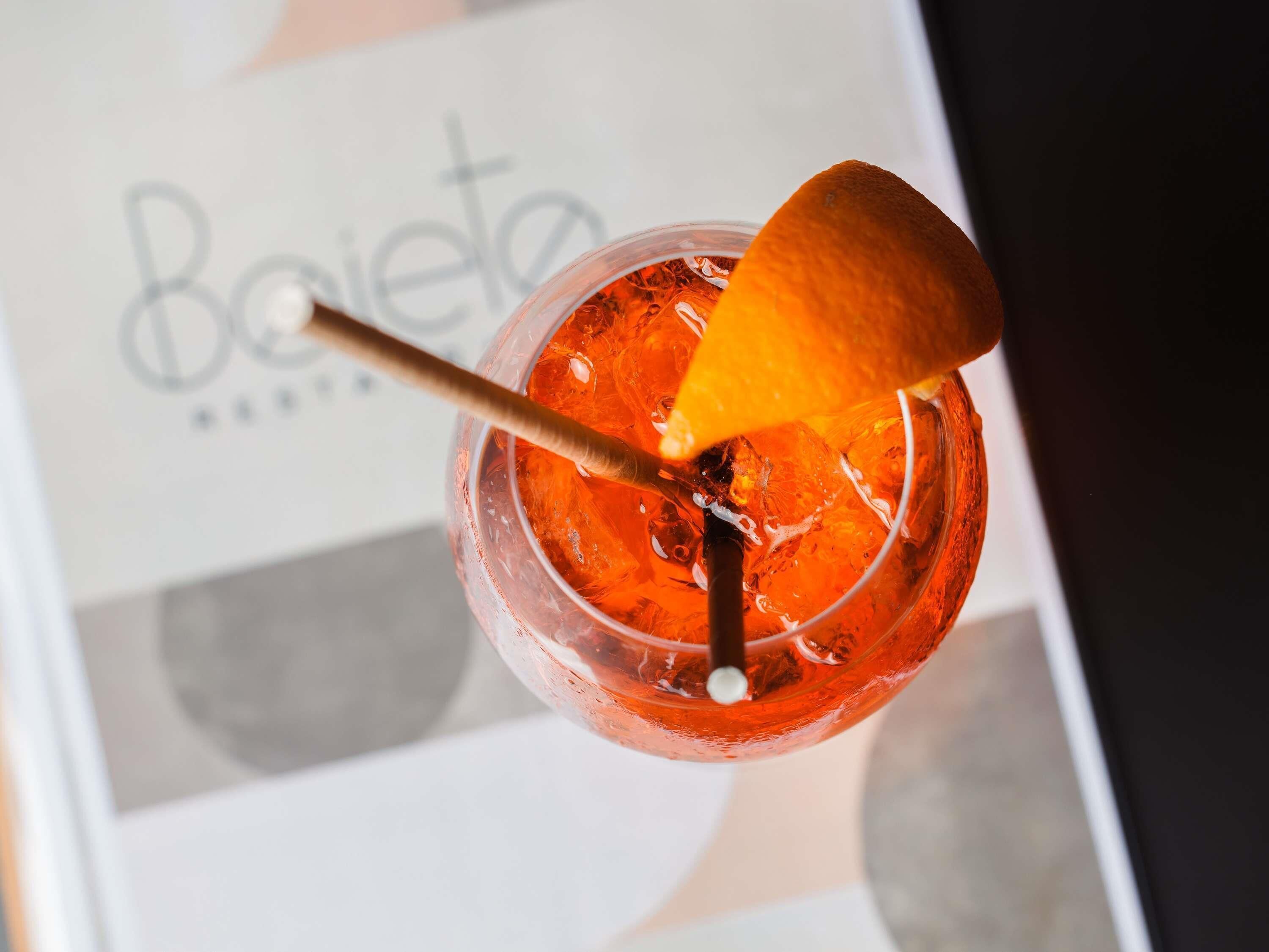 En cocktail med en apelsinskiva som garnering och ett sugrör, med en suddig bakgrund som visar en skylt med ordet ”Boleta”.
