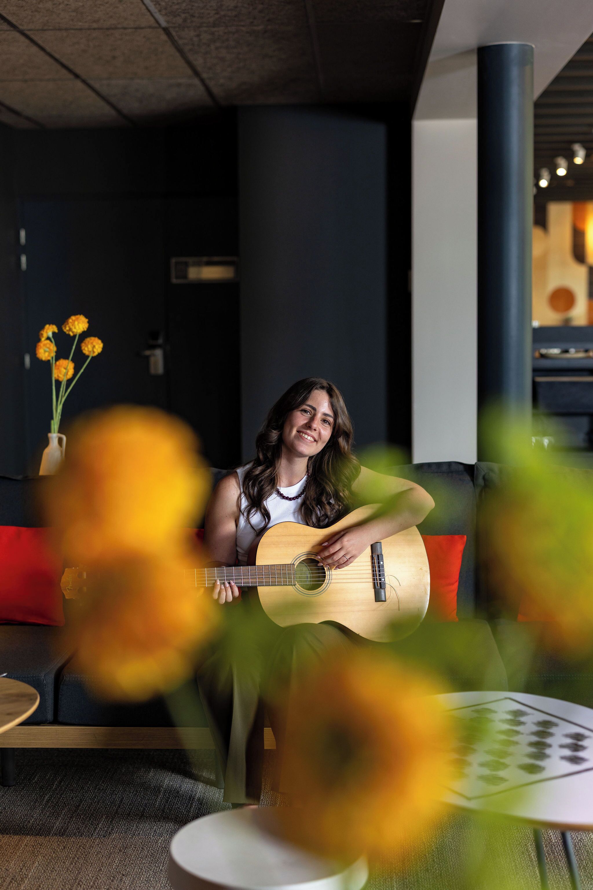 A person playing a guitar in a room with a dark wall, a vase with yellow flowers, and a red cushion.