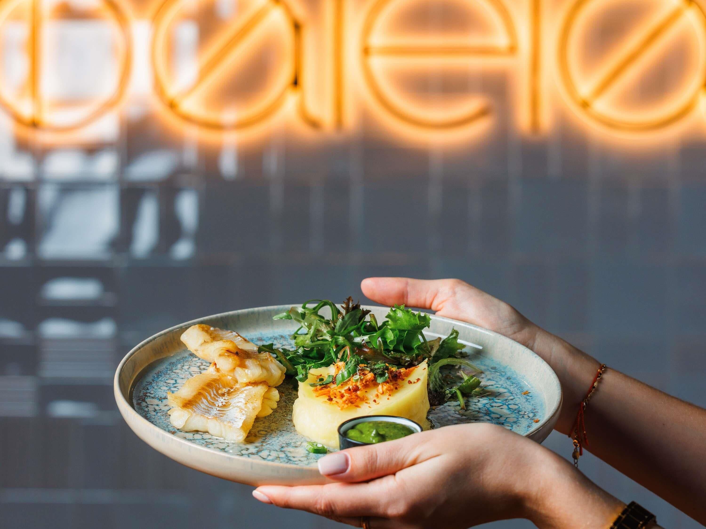 A plate of food with a hand holding it, a neon sign in the background, and a blurred interior.
