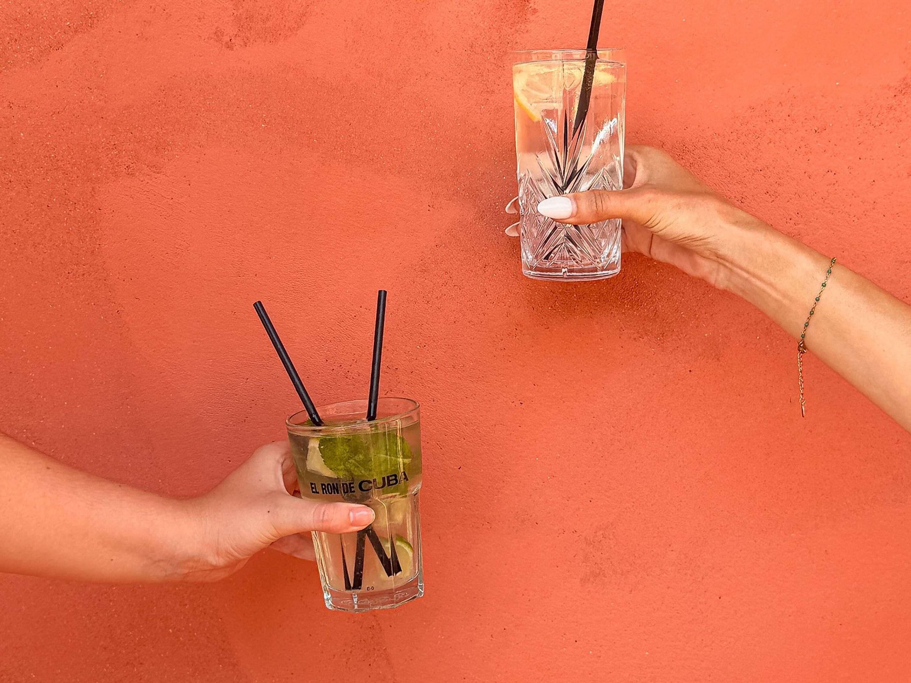 Two hands holding glasses with straws, one labeled 'EL RON DE CUBA' against a textured orange wall.