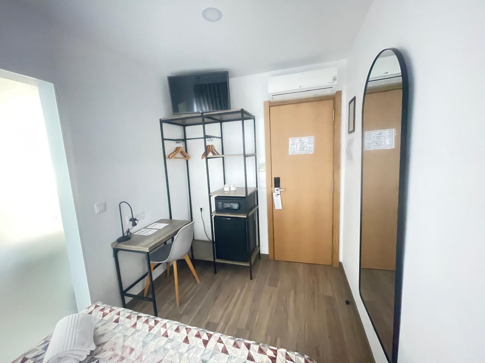 Habitació d'hotel senzilla amb llit, escriptori i cadira, armari, mirall i porta.