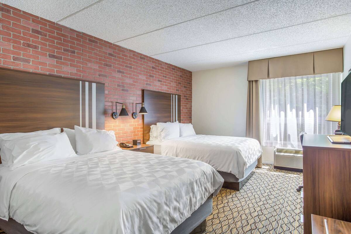 A hotel room with two beds, a brick wall, a wooden headboard, a desk, and a window with curtains.