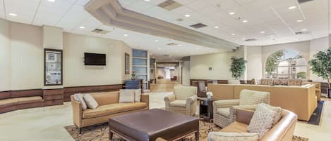 A spacious lobby with multiple seating areas, including sofas and armchairs, a flat-screen TV mounted on the wall, and a large window providing natural light.