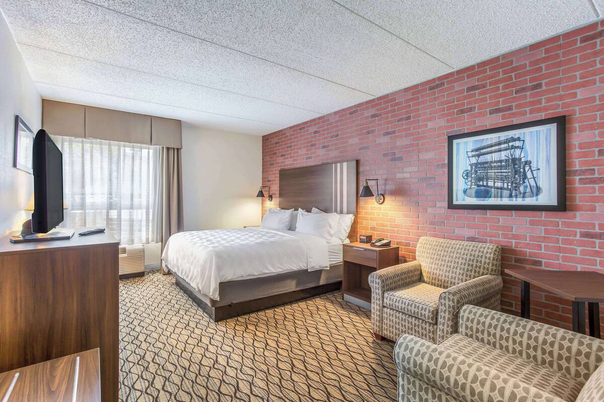 A hotel room with a large bed, a desk with a TV, a seating area with chairs, a brick wall, and a framed picture on the wall.