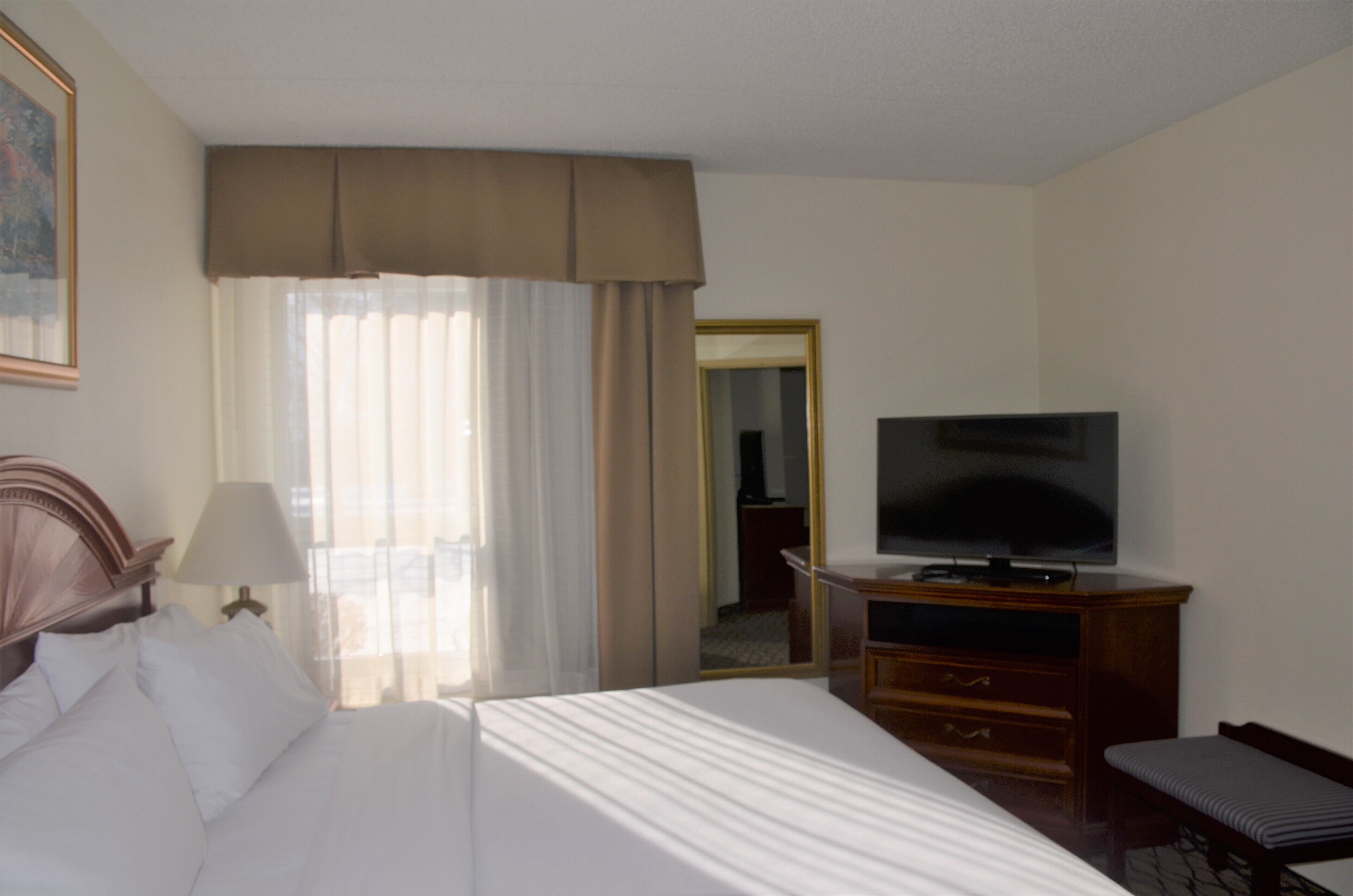 A hotel room with a bed, a TV on a wooden stand, a desk with a lamp, and a window with curtains.