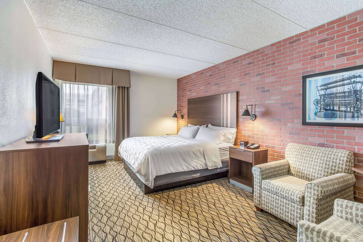 A hotel room with a bed, a TV, a desk, a chair, and a brick wall with a framed picture.