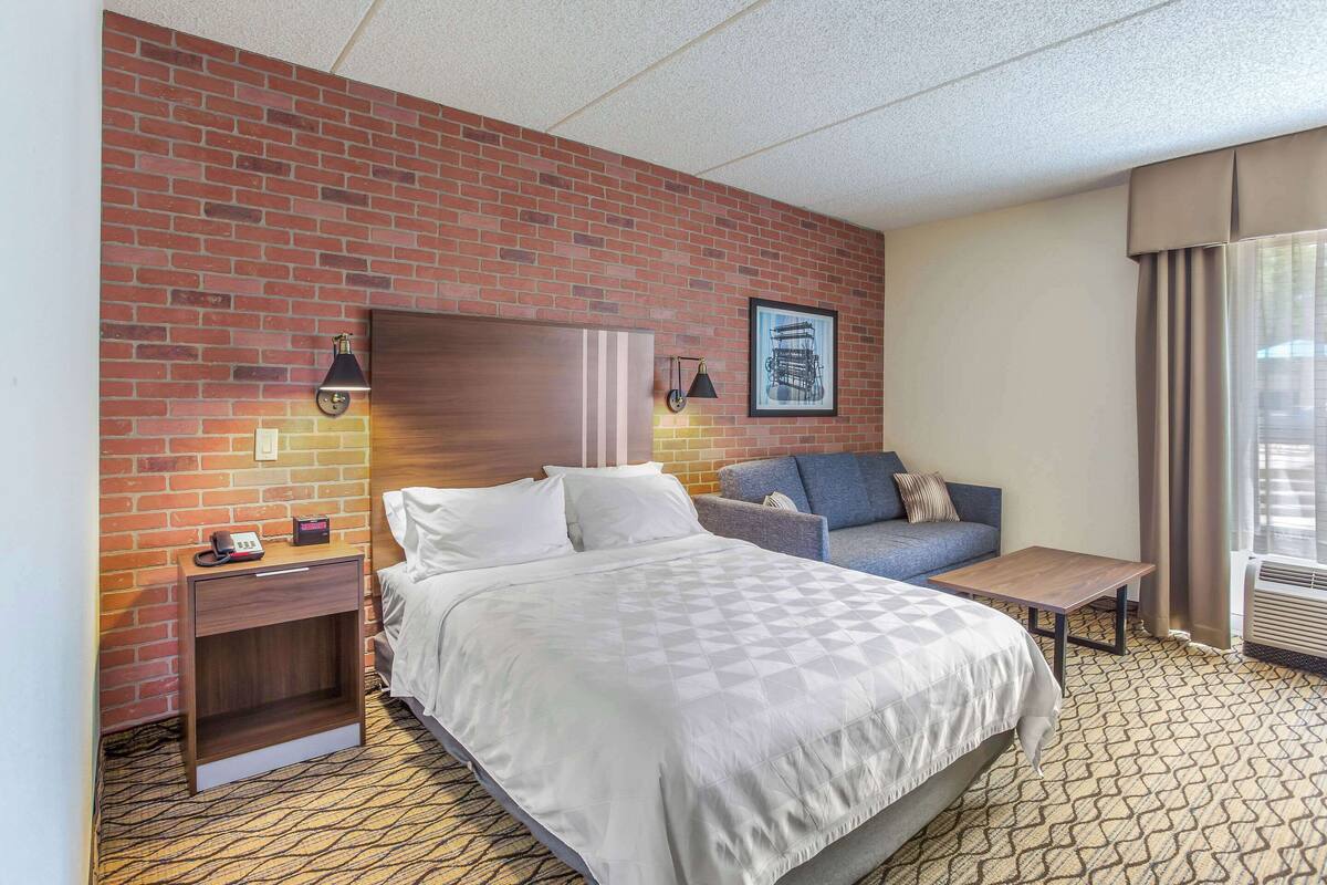 A hotel room with a brick wall, a bed, a sofa, a nightstand, a table, and a picture on the wall.