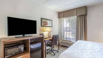 A hotel room with a bed, a desk with a chair, a television, a microwave, a mini-fridge, and a large window with curtains.