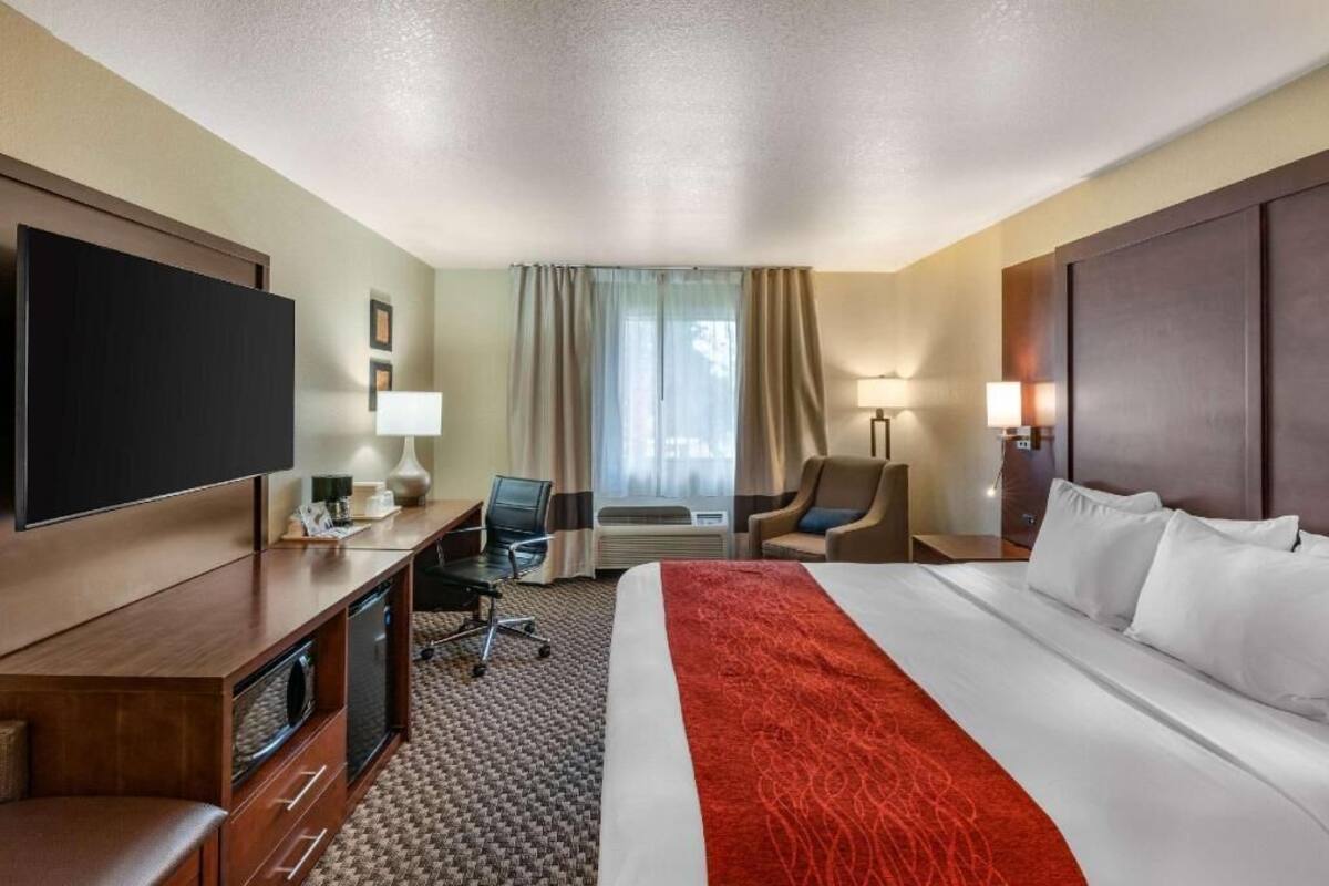 A hotel room with a large bed, a flat-screen TV, a desk with a chair, a lamp, and an air conditioning unit.