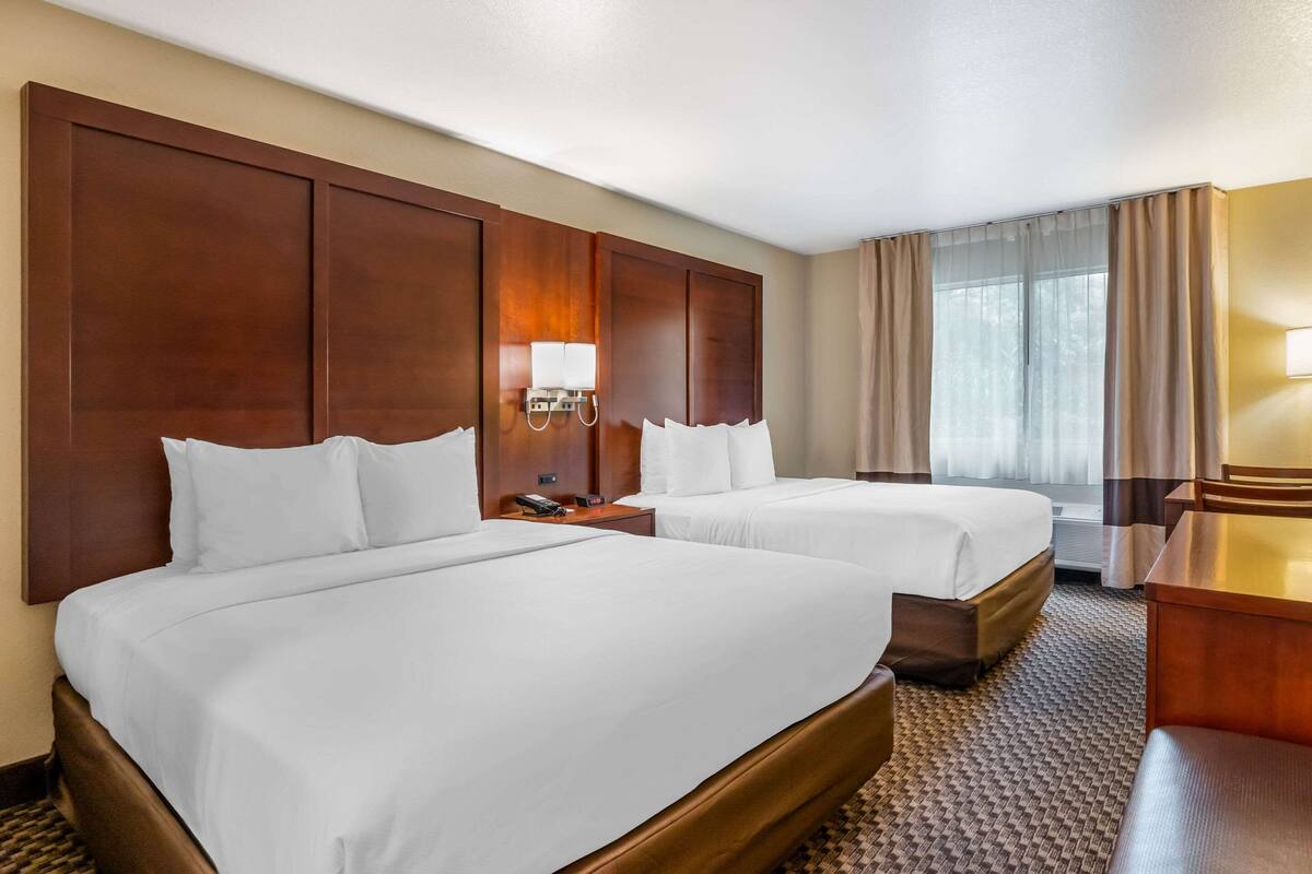 A hotel room with two beds, a desk, a lamp, and a large window with curtains.